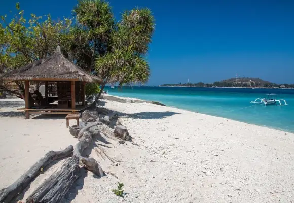 A thatched-roof gazebo on a white sand beach overlooking clear blue water and an outrigger boat in Gili Air.
