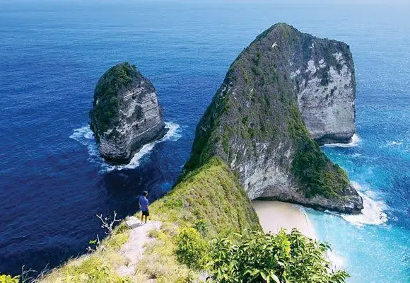 High-angle view of a person standing on the cliff ridge at Kelingking Beach overlooking deep blue ocean waters and the T-Rex headland.