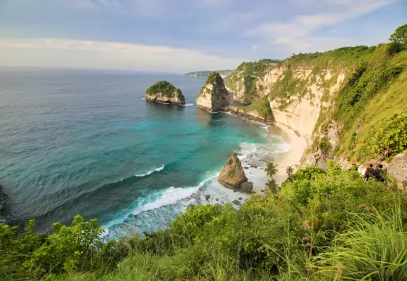 High-angle panoramic view of Atuh Beach showing white sand, turquoise ocean waves, and large rocky islets along the cliffside of Nusa Penida, Bali.