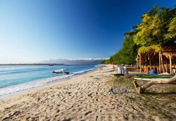 Sunny white sand beach in Gili Trawangan featuring wooden beach shacks, lush trees, and small outrigger boats.